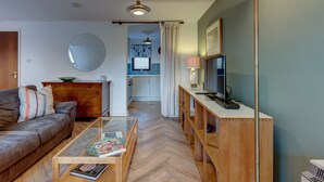 A living room with a sofa, coffee table, and TV stand. A kitchen is visible in the background. - Spires View | Oxford Holiday Let | 1 Bed & Sleeps 2 (Oxford)