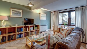 A living room with a sofa, coffee table, bookshelves, a TV, and a window with curtains. - Spires View | Oxford Holiday Let | 1 Bed & Sleeps 2 (Oxford)