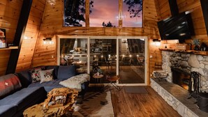 A cozy cabin interior with a wooden interior, a stone fireplace, and a large window overlooking a forest.