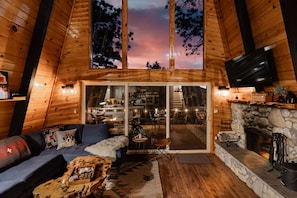 L’intérieur chaleureux d’un chalet, avec un revêtement en bois, une cheminée en pierre et une grande fenêtre donnant sur une forêt.