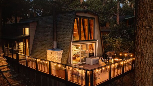 A treehouse with a stone fireplace, a glass door leading to a cozy interior, and a deck with string lights.