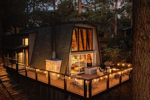 A treehouse with a stone fireplace, a glass door leading to a cozy interior, and a deck with string lights.