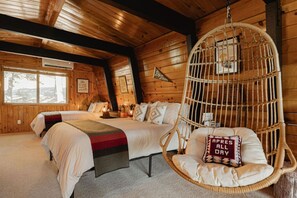 A log cabin room with two beds, a hanging chair, and a window with a view of trees.