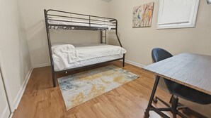 Ein Einzelbett mit Metallgestell, ein kleiner Schreibtisch mit Stuhl, ein Fenster mit Jalousien und ein farbenfrohes abstraktes Gemälde an der Wand.
