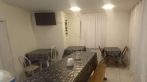 A small, compact room with a single bed, a bedside table, a chair, a desk with a chair, and a television mounted on the wall. - Hostel Canários (Palhoça)