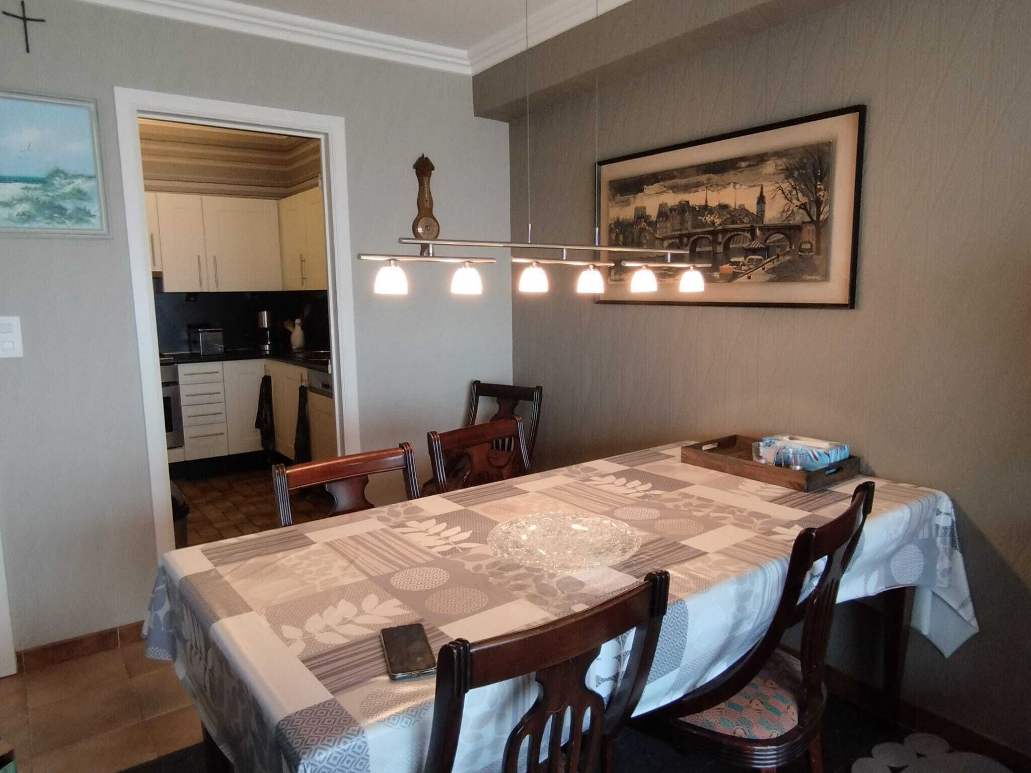 A dining room with a large table, chairs, and a chandelier. There is a kitchen visible in the background.