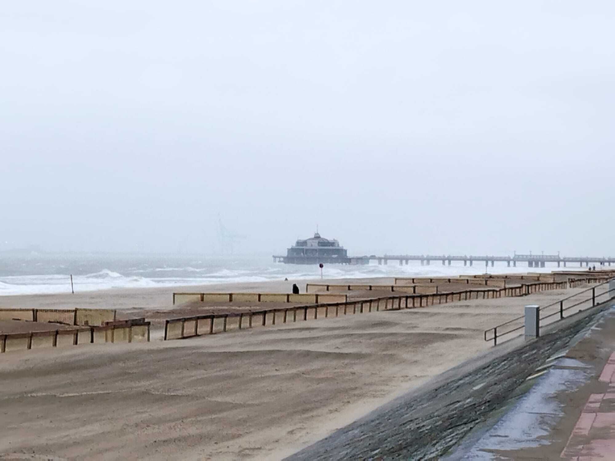 A beach with a long pier extending into the sea, a building at the end of the pier, and a sandy area with a low wall and benches.