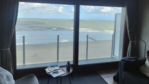 Een hotelkamer met een groot raam met uitzicht op een strand, een tafeltje met een fles en glazen, en een bank.