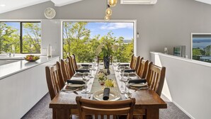 A dining area with a large table set for a meal, a clock on the wall, and a view of trees and sky through large windows. - Ngaio Views - Lake Taupo Holiday Home (Taupo)