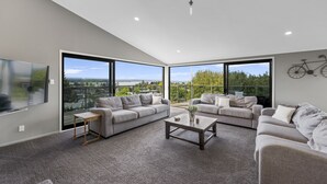 A spacious living room with large windows, grey sofas, a coffee table, and a mounted bicycle. - Ngaio Views - Lake Taupo Holiday Home (Taupo)