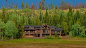 Une grande cabane en rondins à deux étages, dotée de nombreuses fenêtres et d’une cheminée en pierre, entourée d’une forêt dense et d’un champ herbeux.
