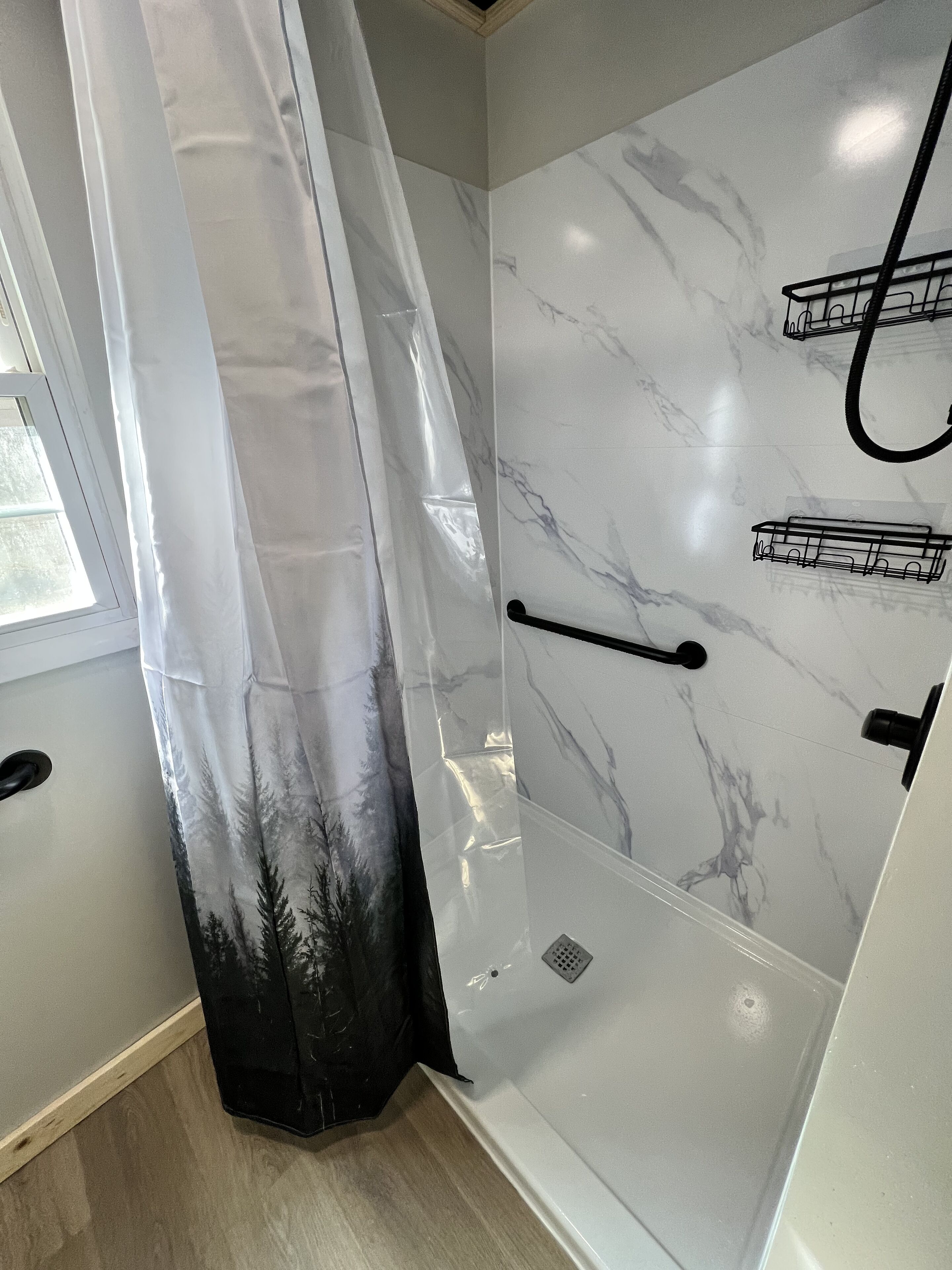 A modern bathroom with a glass shower enclosure, marble walls, and a window with a view of trees.