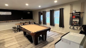 Une table de billard dans une pièce dont le sol est en bois, avec un ventilateur de plafond et un climatiseur fixé au mur.