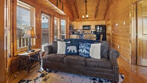 L’intérieur chaleureux d’un chalet avec un plafond en bois, un canapé en cuir marron agrémenté de coussins décoratifs, une table à manger avec une lampe et un coin cuisine équipé d’appareils électroménagers.