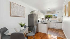 A modern kitchen with a central island, white cabinetry, and a dining area with a glass table and grey chairs. - Broadway by The Beach (Glenelg South)