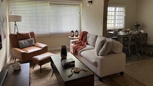 A living room with a brown leather armchair, a white sofa, a coffee table with books and a vase, a dining area with a table and chairs, and a large window with blinds. - Island Oasis, Near Magens Bay Beach with Back-Up Power System (Charlotte Amalie)