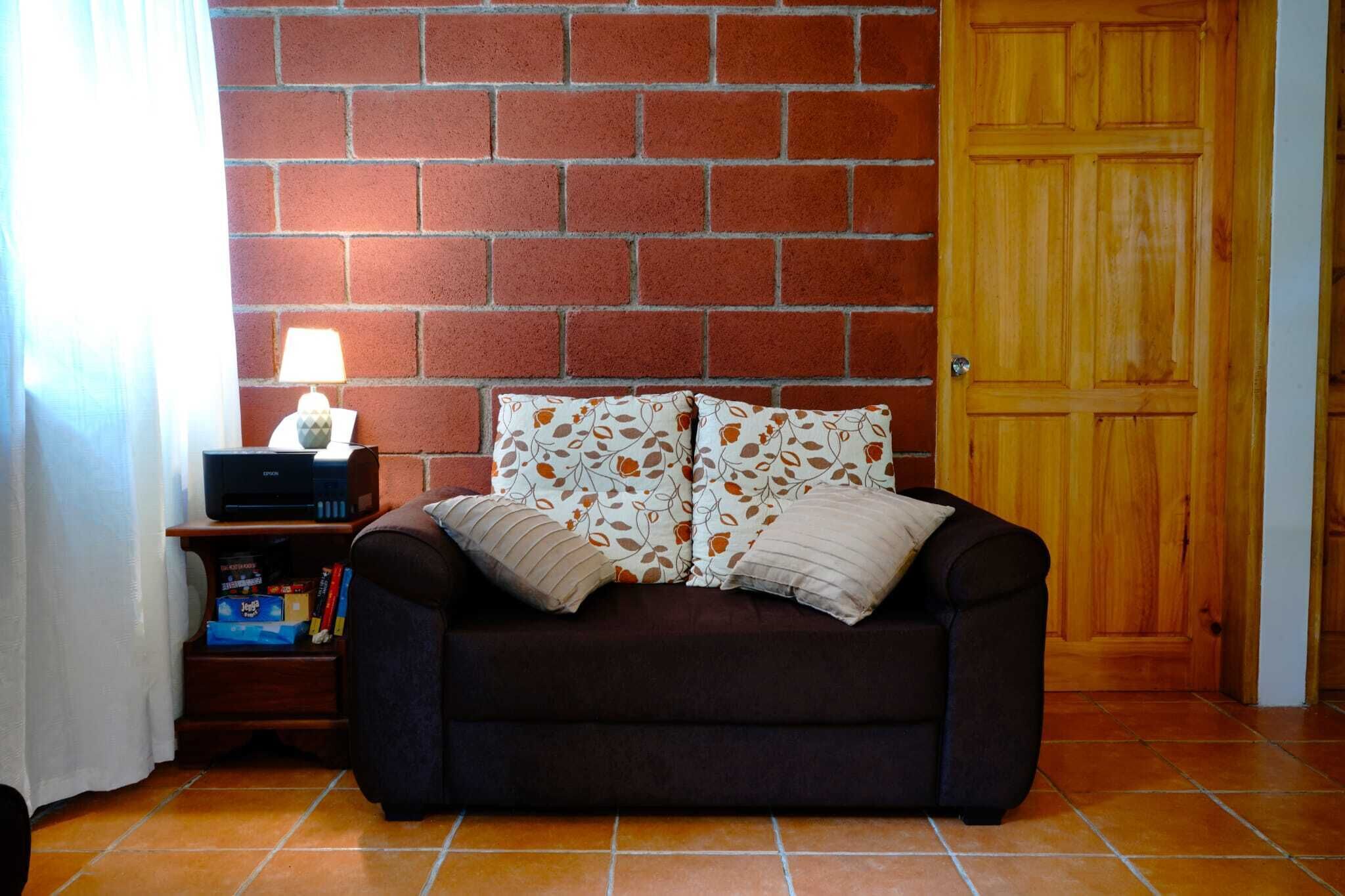 A cozy room with a brick wall, a wooden door, a dark brown sofa with patterned cushions, a small wooden side table with a lamp, and a shelf with books and a box.