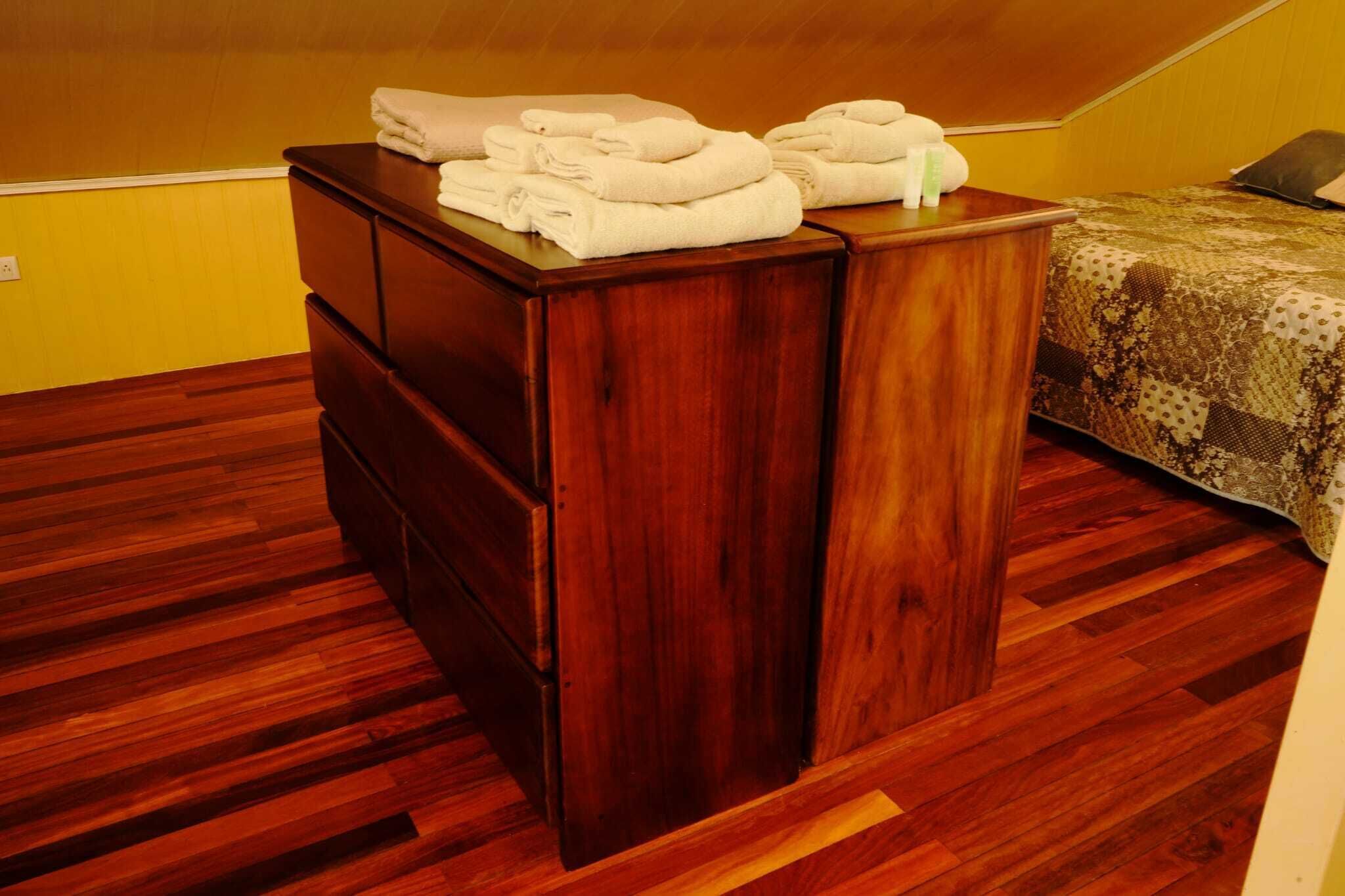 Uma cômoda de madeira com toalhas dobradas e artigos de higiene pessoal em um quarto com piso de madeira e uma cama com colcha estampada.