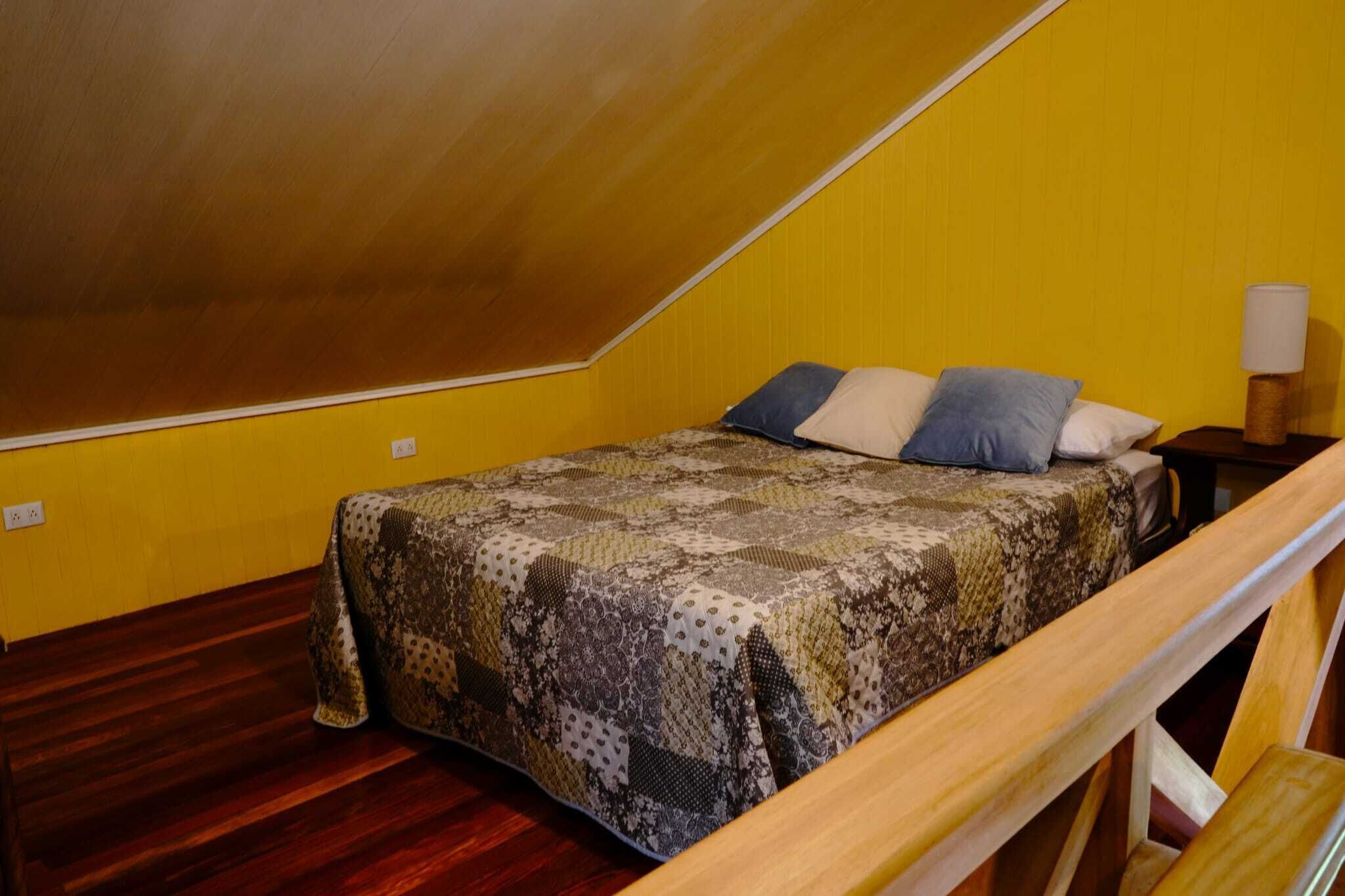 A small, triangular attic bedroom with a wooden ceiling and yellow walls. It features a bed with a patterned quilt and several pillows, a nightstand with a lamp, and a wooden bench.