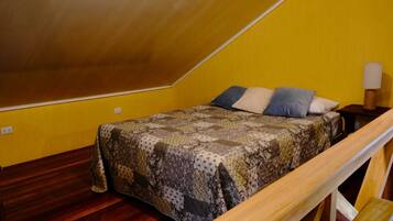A small, triangular attic bedroom with a wooden ceiling and yellow walls. It features a bed with a patterned quilt and several pillows, a nightstand with a lamp, and a wooden bench.