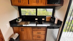 Een kleine keuken met houten kastjes, een zwart aanrecht, een roestvrijstalen koelkast en een raam met uitzicht op groen.