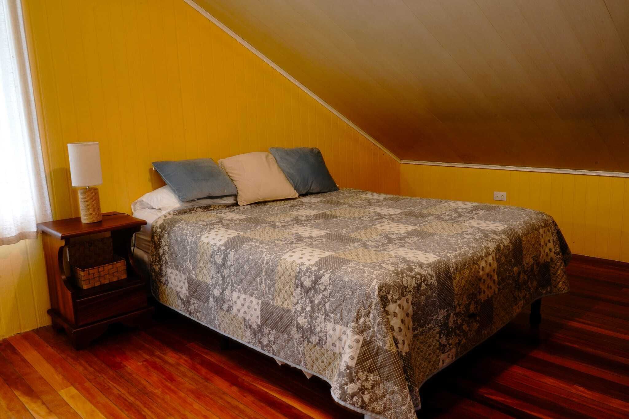 A bedroom with a sloped ceiling, a bed with patterned bedding, a nightstand with a lamp, and wooden flooring.