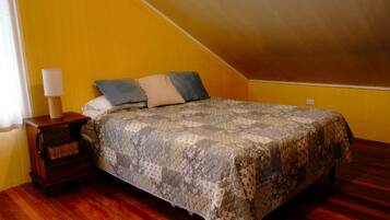 A bedroom with a sloped ceiling, a bed with patterned bedding, a nightstand with a lamp, and wooden flooring.