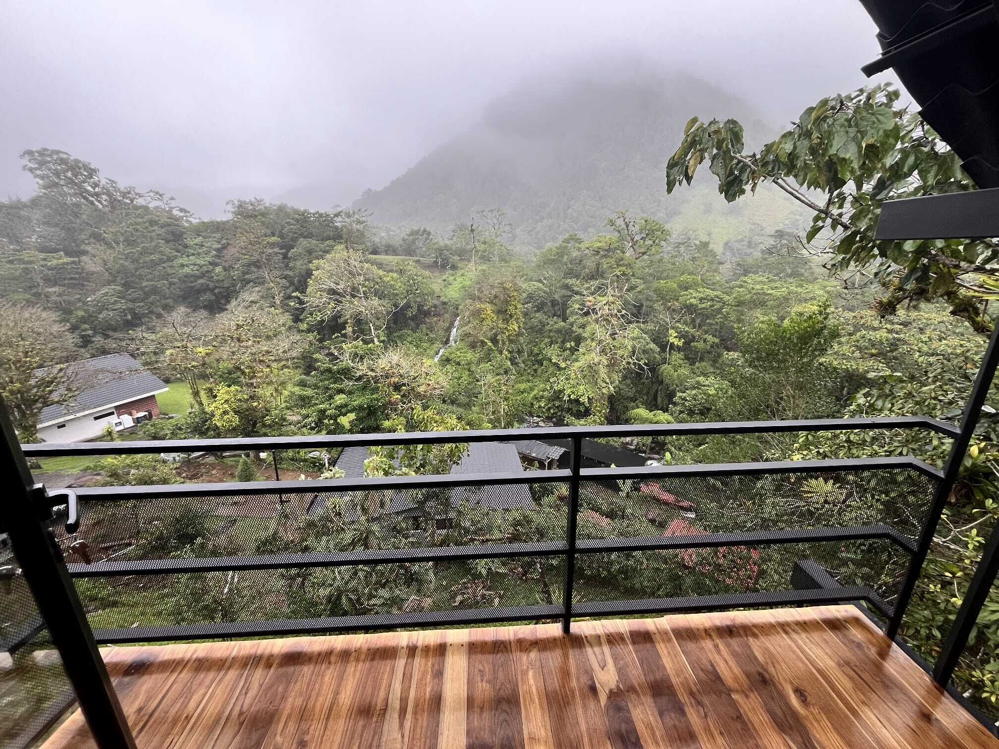 Un balcón con suelo de madera y baranda de metal que da a un paisaje verde exuberante con una cascada.