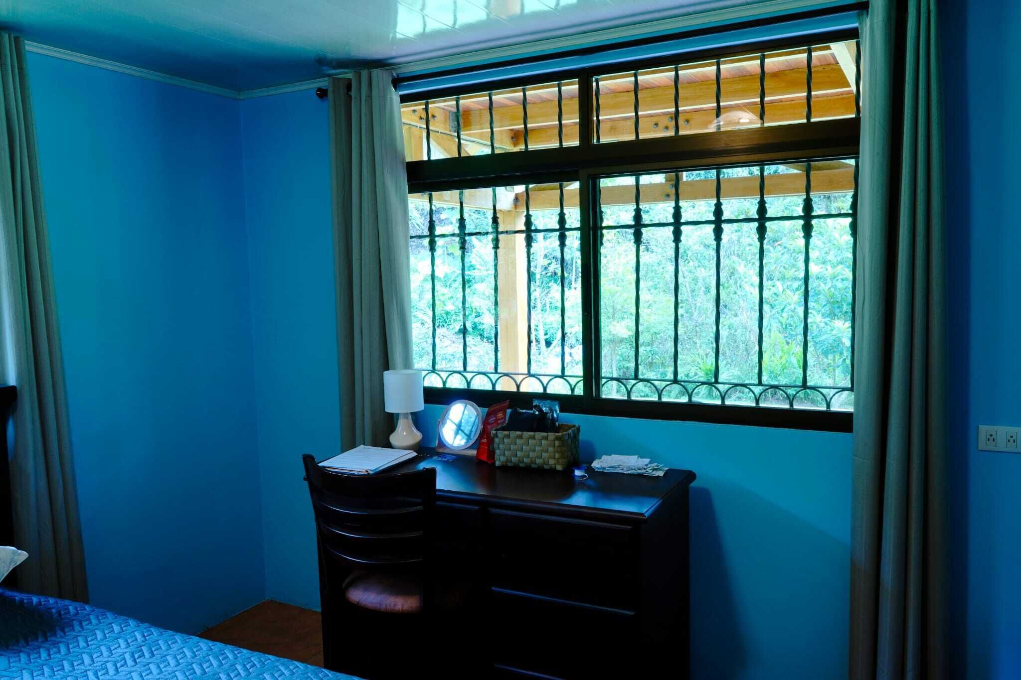 A room with a desk, a lamp, a clock, and a window with a view of greenery.
