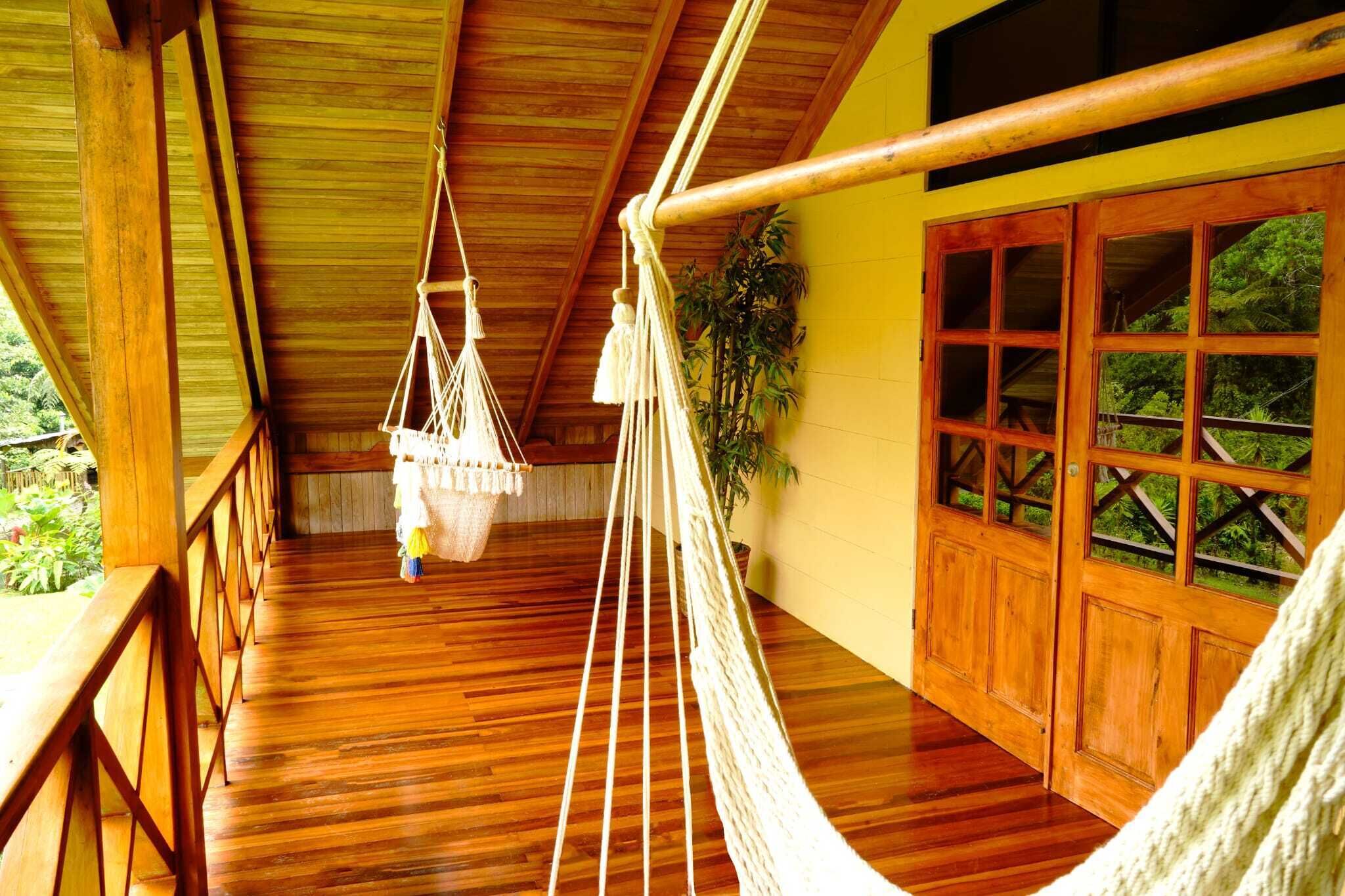 A wooden balcony with a hammock and a hanging chair, wooden floors, and a view of greenery outside.