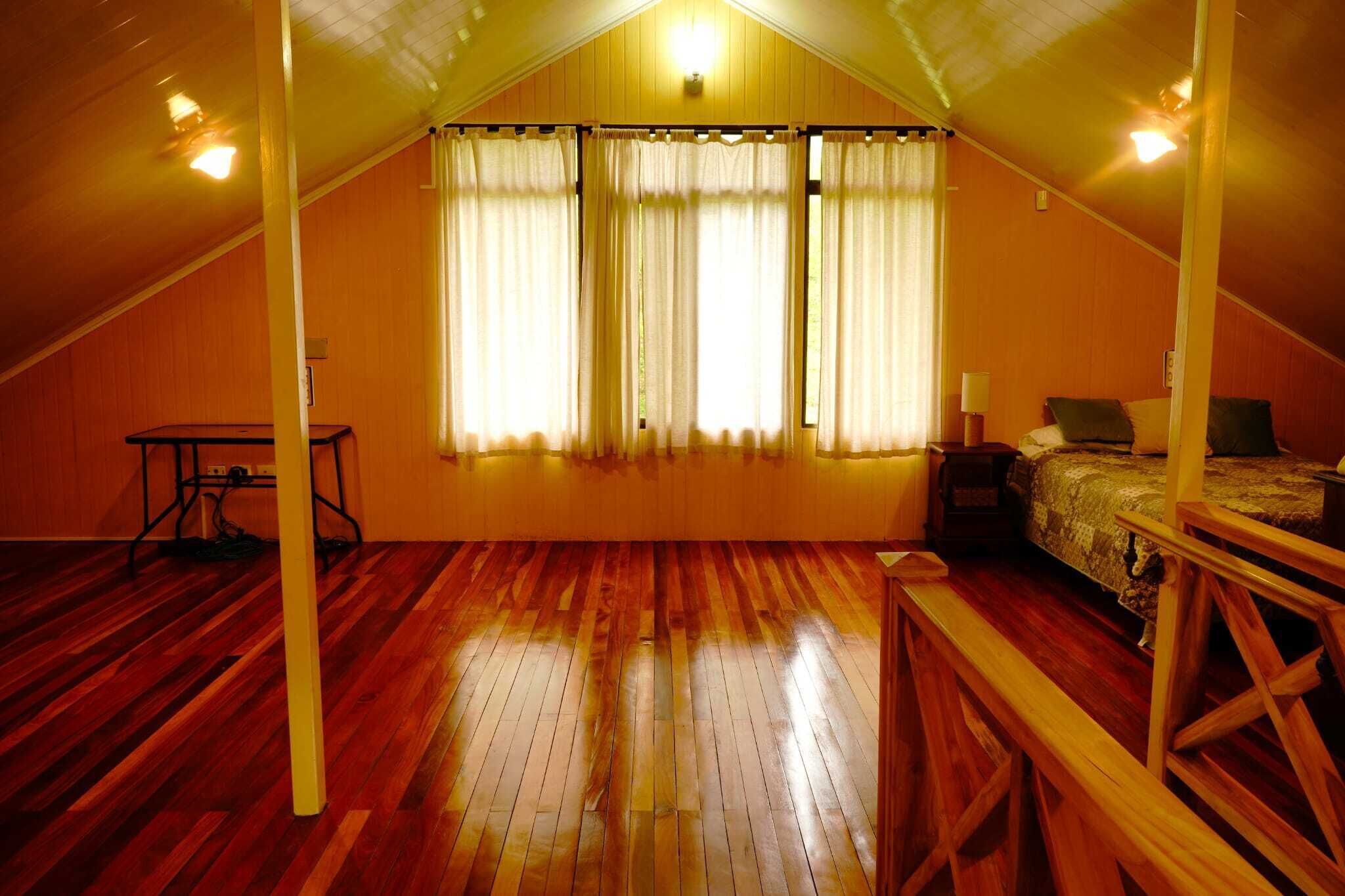 A room with a sloped ceiling, wooden flooring, a bed with a patterned bedspread, a bedside table with a lamp, and a window with curtains.