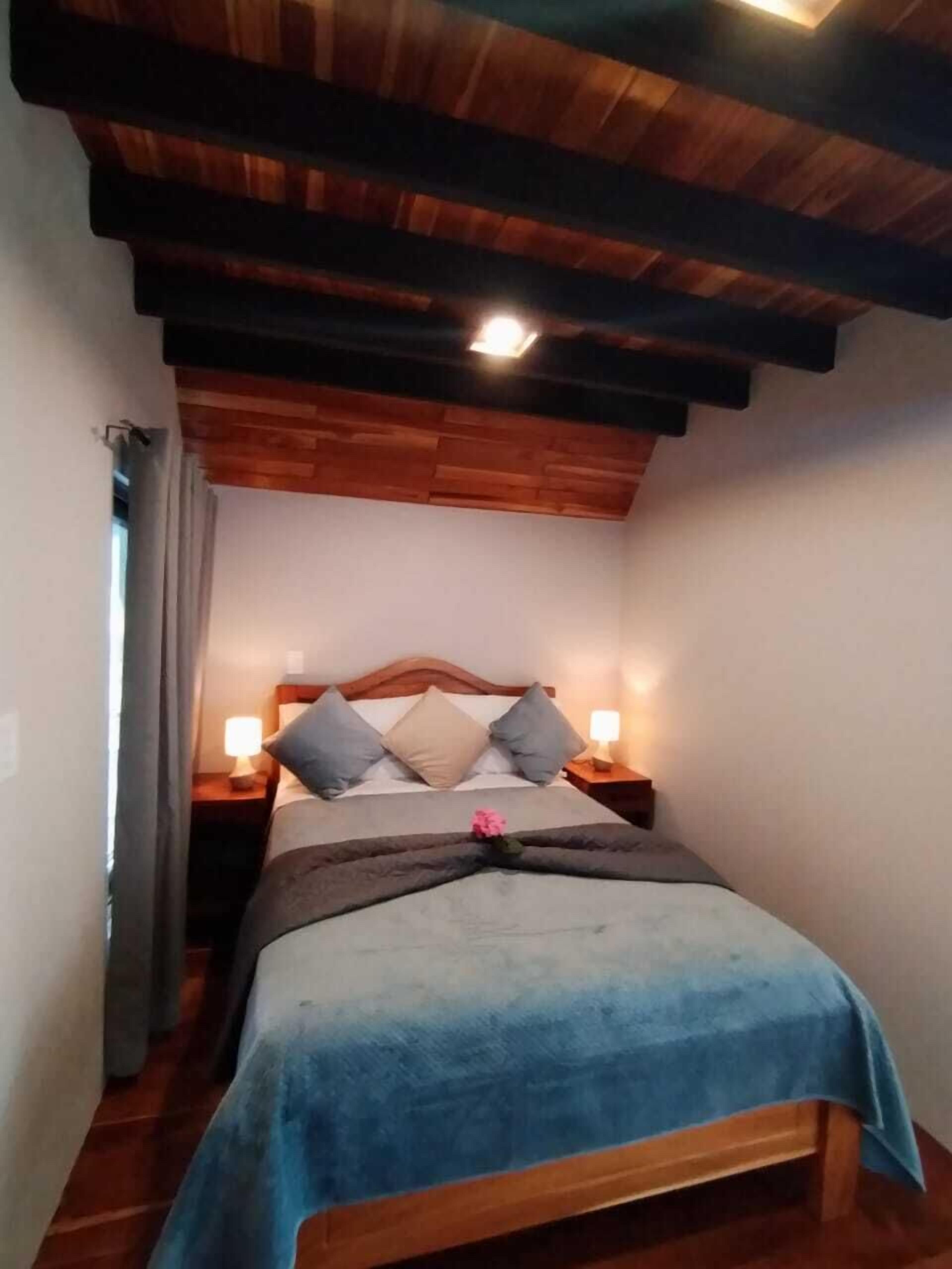 Un dormitorio con techo de madera, una cama con manta azul, dos lámparas de noche y una mesita de noche.