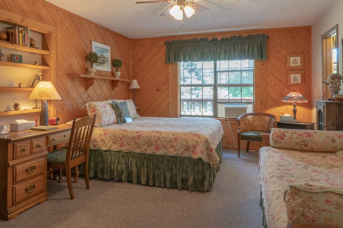 A cozy bedroom with a wooden paneled wall, a large bed with floral bedding, a desk with a lamp, and a window with a view of trees.