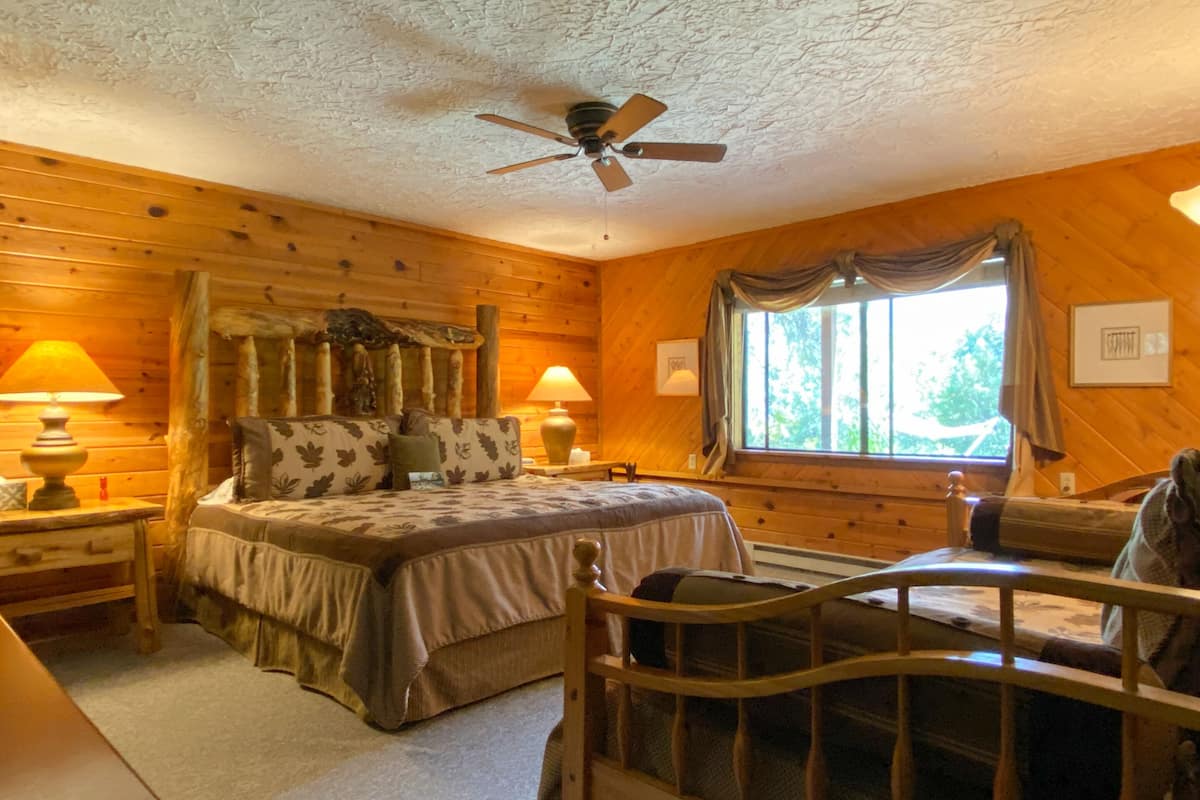 A cozy log cabin bedroom with a bed, a chair, a nightstand, a lamp, a ceiling fan, and a window with curtains.