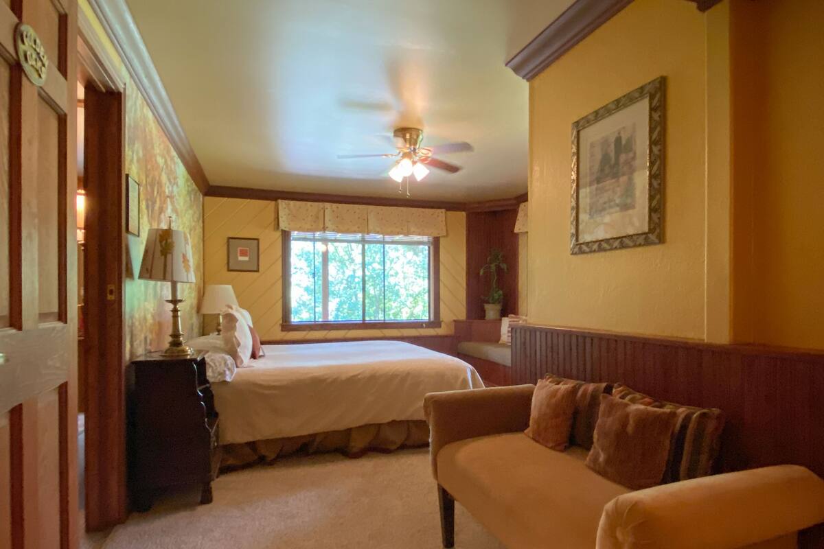 A bedroom with a double bed, a nightstand, a lamp, a window with a view of trees, a ceiling fan, and a framed picture on the wall.