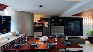 Uma zona de refeições moderna com mesa de madeira posta para uma refeição, luminária pendente e cozinha planejada com frigorífico e prateleiras.