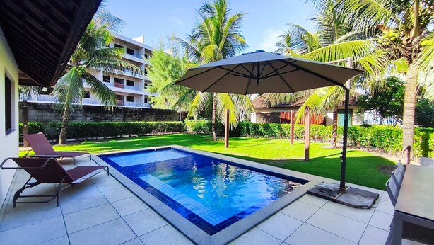 A swimming pool with lounge chairs, a covered area with an umbrella, and surrounding greenery. - Lindos Bangalôs - Carneiros Beach Resort (Tamandaré)