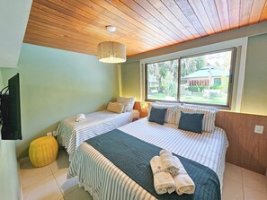 A hotel room with two beds, a window with a view of the outdoors, a ceiling fan, and a television. - Lindos Bangalôs - Carneiros Beach Resort (Tamandaré)