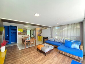 A modern living room with a sofa, ottoman, dining table, chairs, and a kitchen area in the background. - Lindos Bangalôs - Carneiros Beach Resort (Tamandaré)