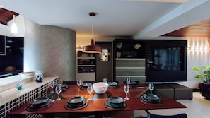 A modern dining area with a wooden table set for a meal, a hanging pendant light, and a built-in kitchen with a refrigerator and shelves. - Lindos Bangalôs - Carneiros Beach Resort (Tamandaré)