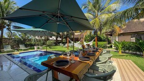 A poolside dining area with a table set for four, surrounded by chairs, under a large umbrella. - Lindos Bangalôs - Carneiros Beach Resort (Tamandaré)
