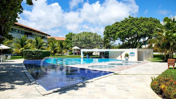 A swimming pool with a tiled deck, surrounded by palm trees and a building with a red roof. - Lindos Bangalôs - Carneiros Beach Resort (Tamandaré)