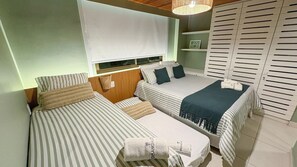 A compact room with two single beds, a small bedside table, a wall-mounted shelf, and a window with blinds. - Lindos Bangalôs - Carneiros Beach Resort (Tamandaré)
