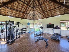 A gym with exercise equipment, a bench, a treadmill, and a TV on the wall. - Lindos Bangalôs - Carneiros Beach Resort (Tamandaré)