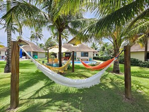 A hammock between palm trees in a well-maintained garden with a pool and a house in the background. - Lindos Bangalôs - Carneiros Beach Resort (Tamandaré)