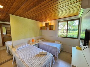 A room with two beds, a small table, a window with a view of greenery, and a wooden ceiling. - Lindos Bangalôs - Carneiros Beach Resort (Tamandaré)