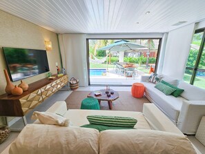 A modern living room with a flat-screen TV, a sofa, a coffee table, and a view of an outdoor patio with a pool. - Lindos Bangalôs - Carneiros Beach Resort (Tamandaré)