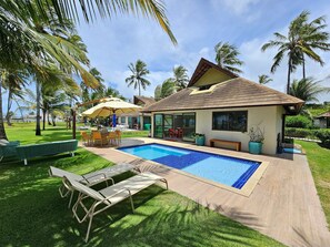 A poolside area with lounge chairs, a pool, and a house with a tiled roof. - Lindos Bangalôs - Carneiros Beach Resort (Tamandaré)