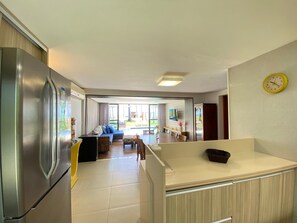 A modern kitchen with a refrigerator, a dining area with a table and chairs, and a view of the outdoor area. - Lindos Bangalôs - Carneiros Beach Resort (Tamandaré)