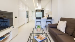 Una sala de estar moderna con una mesa de centro de vidrio, un sofá marrón y una zona de cocina al fondo.